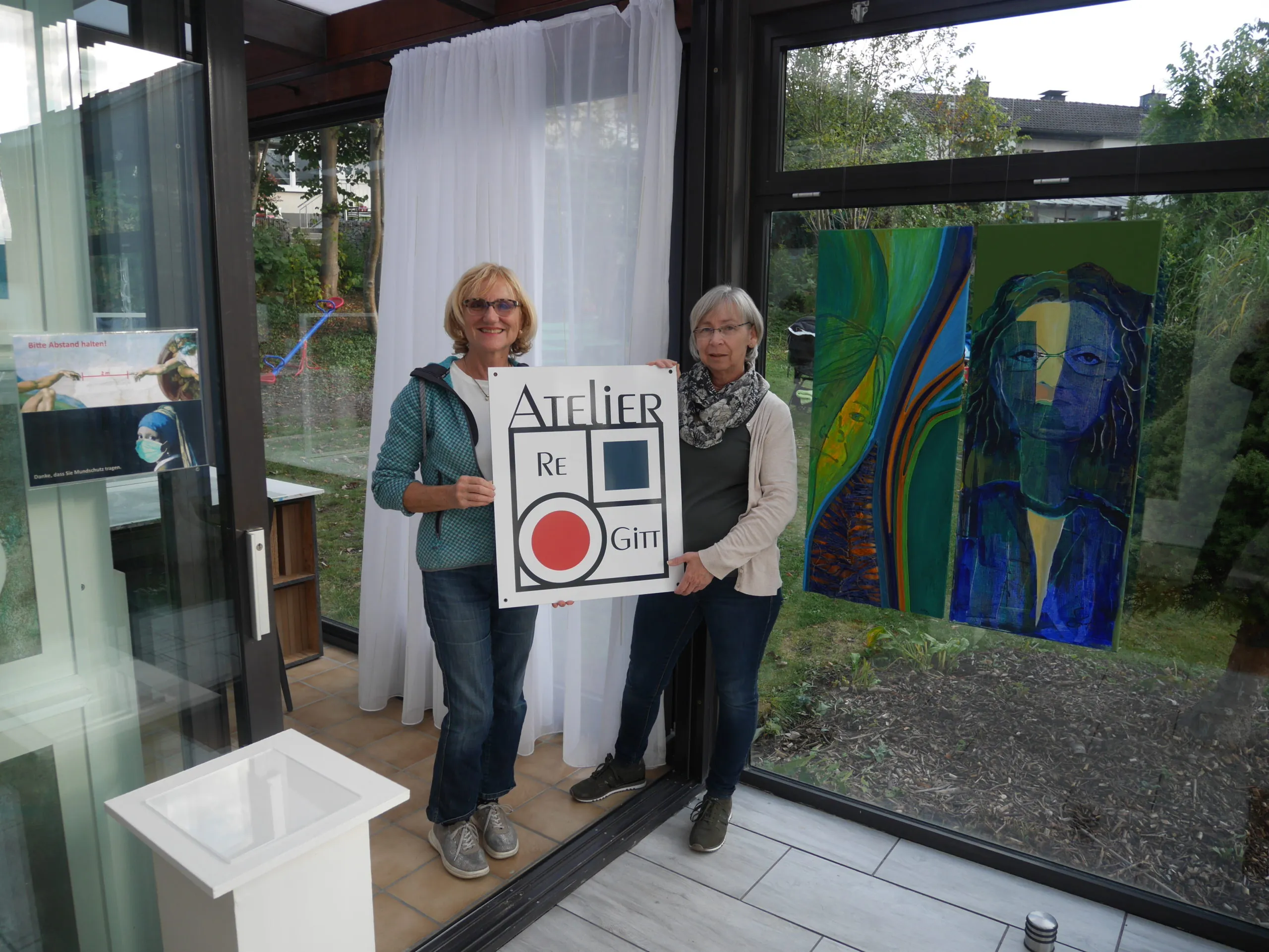 Zwei Frauen halten das Schild des Ateliers ReGitt, im Hintergrund farbige Gemälde